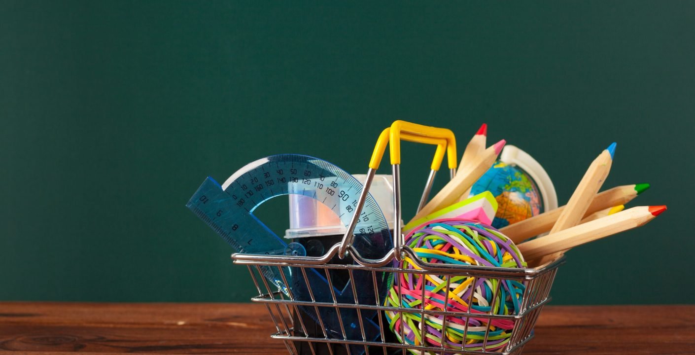 Panier de fournitures scolaires (crayons de couleur, règle, rapporteur, petit globe) devant un tableau vert